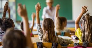 Rear view of large group of students raising their arms to answer the question on a class at elementary school.